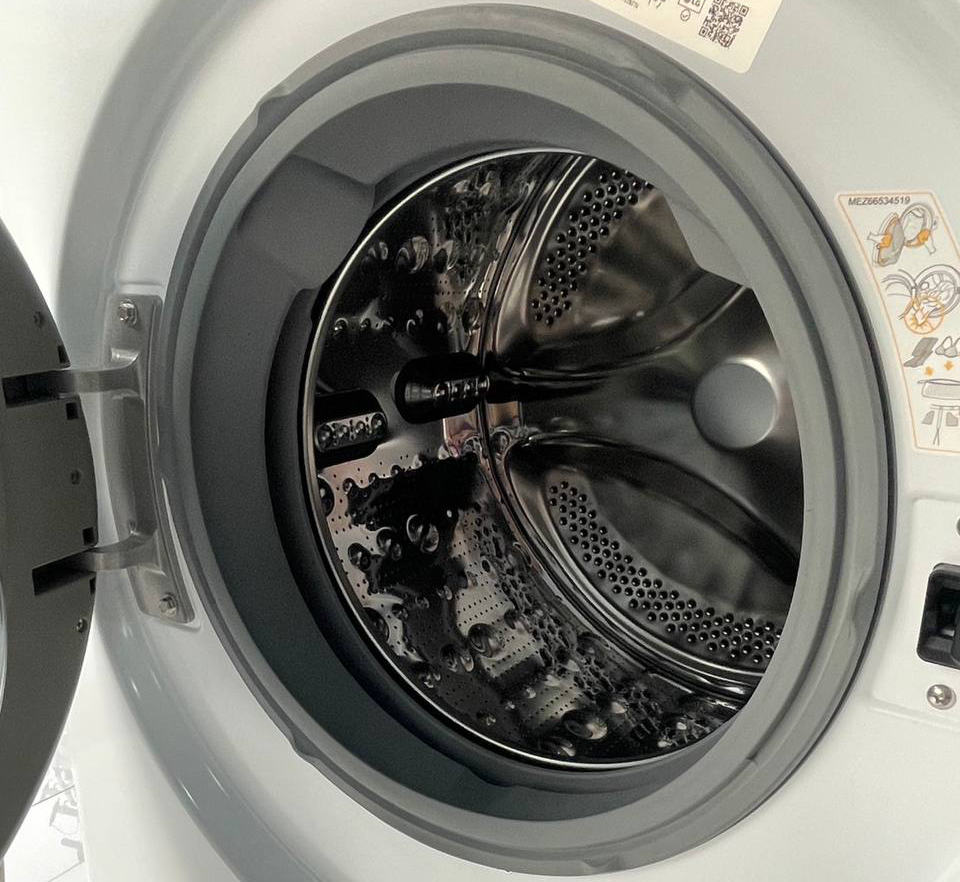 A hand is placing or removing a blue towel from a front-loading washing machine. The machine door is open, revealing additional clothes ready to wash or dry. The surroundings are partially visible, suggesting a laundry setting.