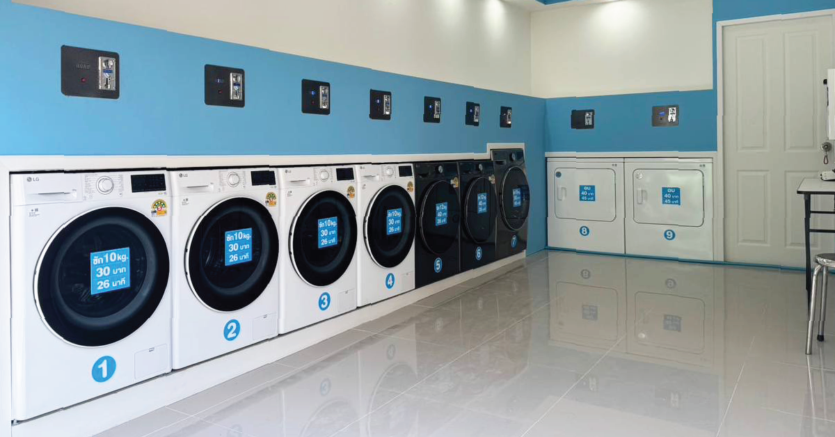 A row of washing machines against a blue and white wall, each with a digital display and numbered 1 to 7. Payment devices are mounted on the wall above each machine.